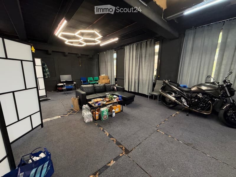 A spacious, modern room with a grey carpeted floor, a black sofa, a small table with snacks, a screen, and a motorcycle parked in the corner.