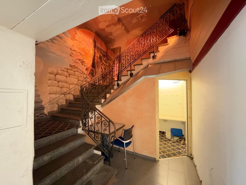 Hallway with a staircase featuring ornate metal railings, a mural on one wall, and a small blue chair near the stairs.