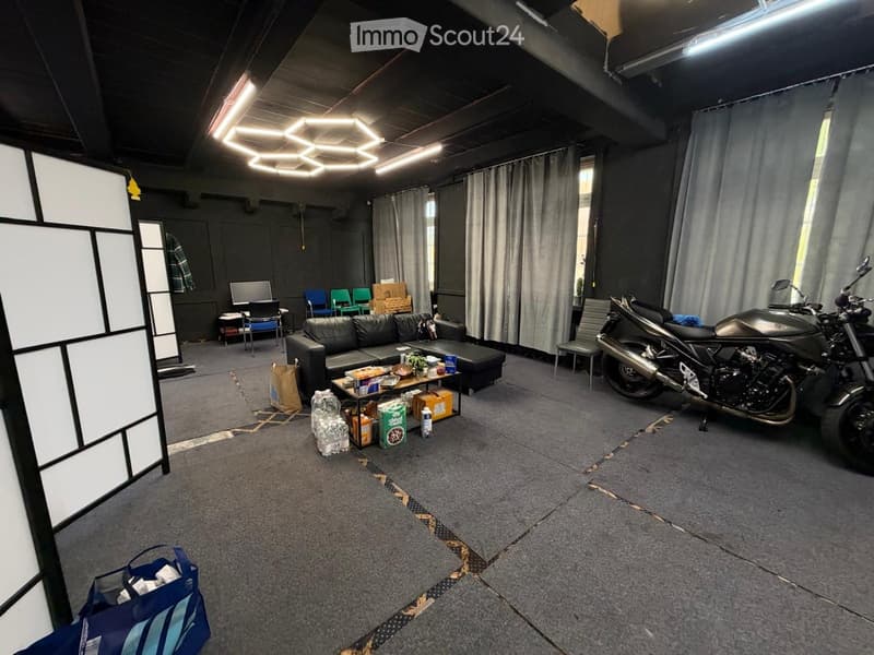 A spacious room with a grey carpeted floor, a black leather sofa, a small table with snacks, a motorcycle, and several chairs arranged around the space.