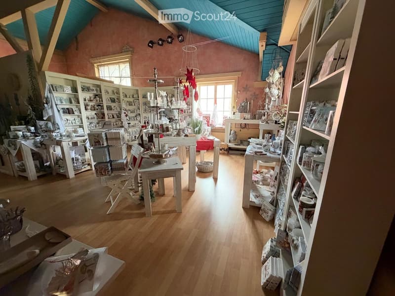 A cozy, well-lit room with wooden flooring, white shelves filled with decorative items, a small table with a candle holder, and a hanging star decoration. The room features a slanted ceiling with exposed wooden beams and warm lighting.