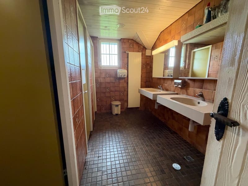 Bathroom with two sinks, a mirror, a cabinet, and a trash can. The walls are tiled in a warm brown color, and the floor is tiled in a dark brown color.
