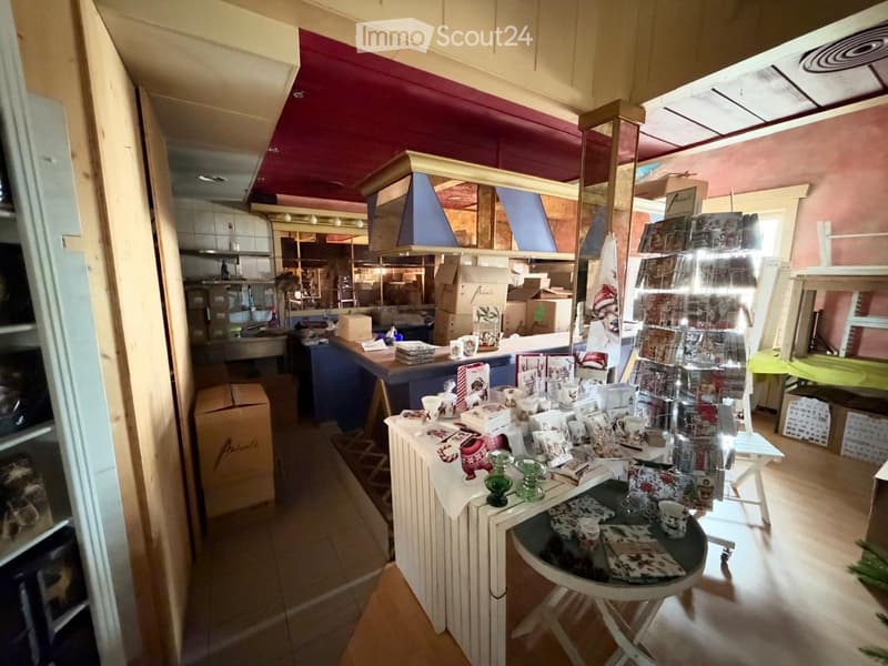 A small shop or store with a counter, display racks, and a kitchen area in the background. The counter has cups, a small table, and decorative items.