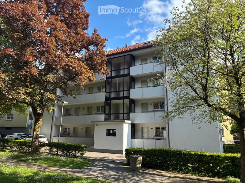 Modern multi-story apartment building with white facade, large windows, and balconies, surrounded by trees and greenery.
