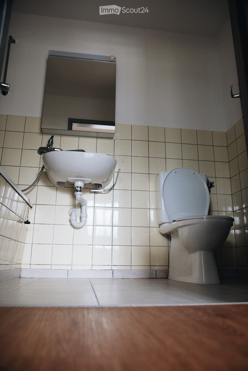 A small bathroom with a toilet, sink, and mirror. The walls are tiled, and the floor is a combination of tiles and wood.