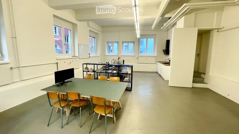 A room with a green table surrounded by four chairs, a shelf with books, a screen, and a small sink area.