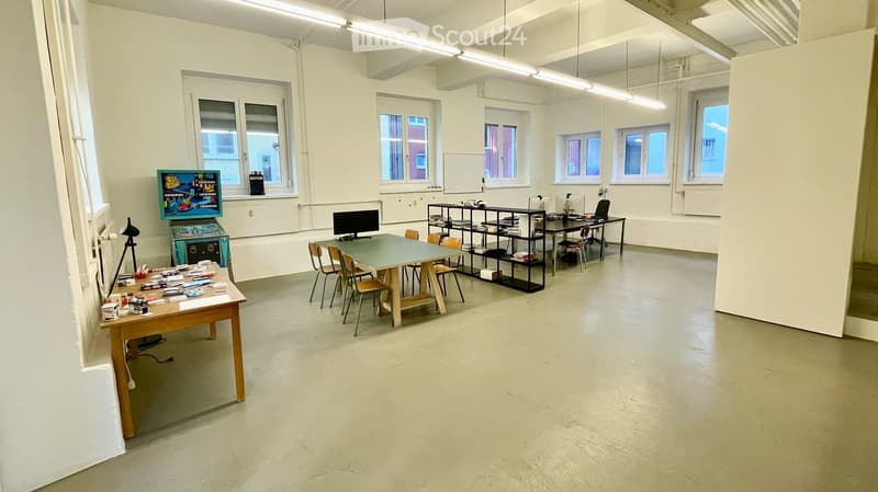 A spacious, well-lit room with multiple tables, chairs, shelves, a screen, and an arcade machine. The room has large windows and a clean, minimalist design.