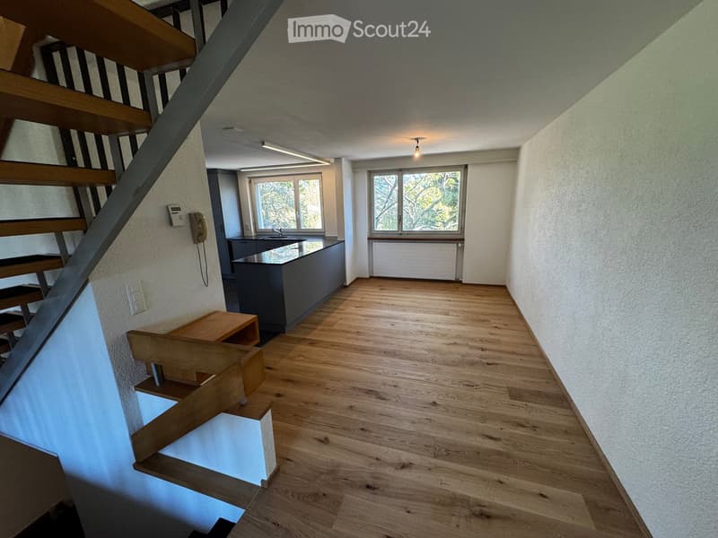 Empty room with wooden flooring, a staircase leading upwards, and a small kitchen area with a sink and countertop. The walls are painted in a light color, and there are windows allowing natural light to enter.
