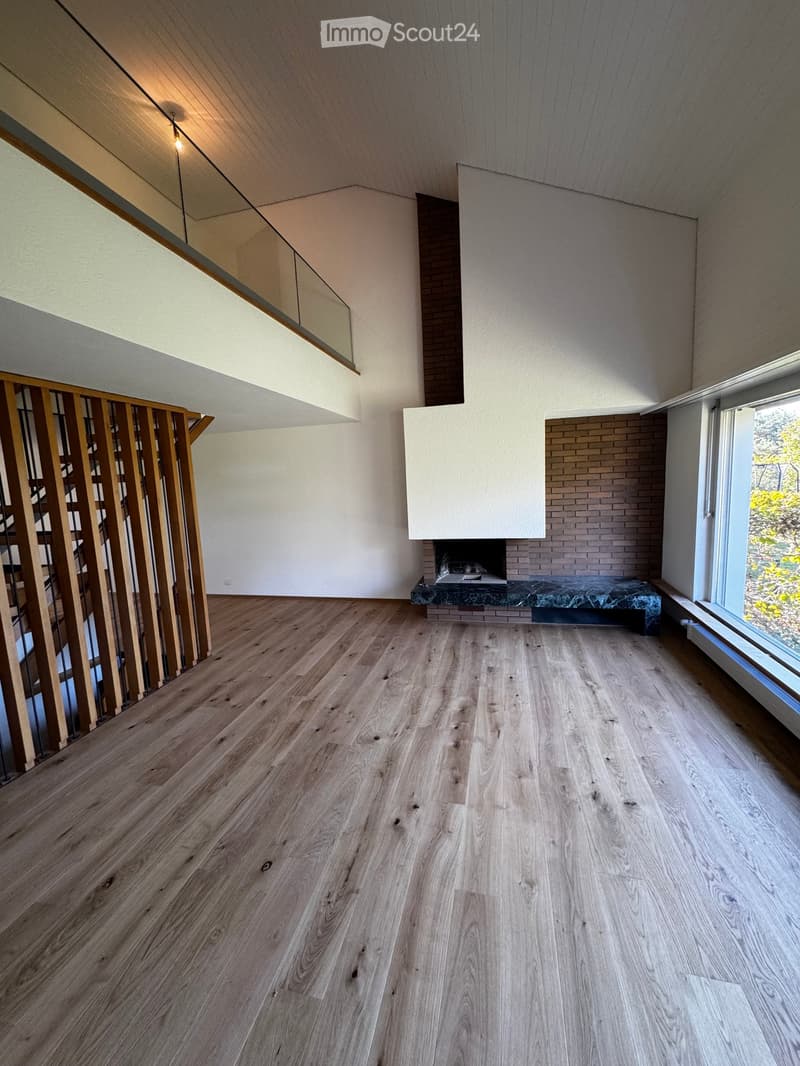 Empty room with wooden flooring, white walls, and a fireplace with a marble surround. Large windows provide natural light, and a wooden railing is visible on the left side.