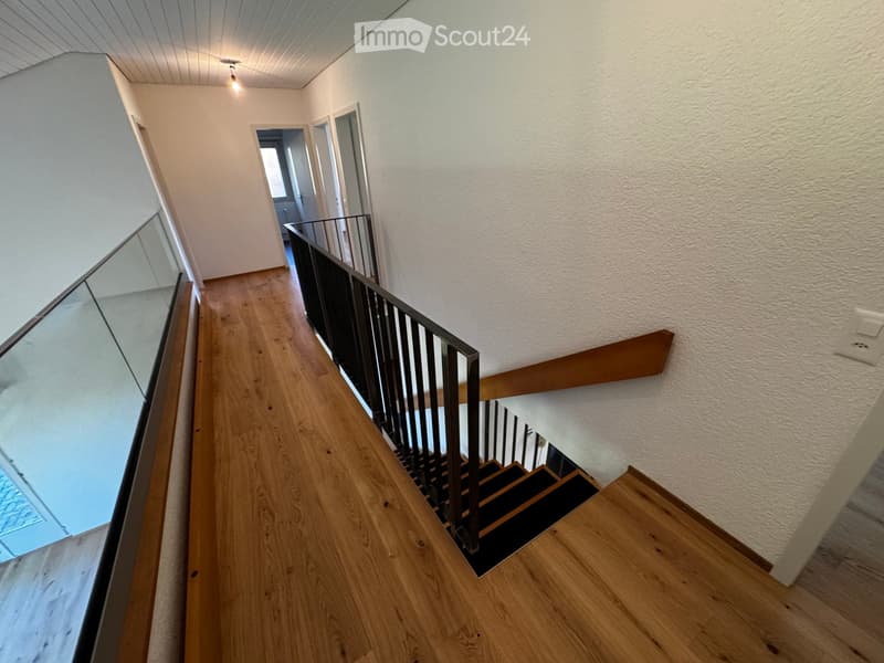 Modern hallway with wooden flooring, white walls, and a staircase with a metal railing and glass side.