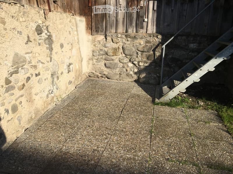 Raised concrete ground floor area adjacent to a stone wall, with a metal staircase leading up to a wooden structure.