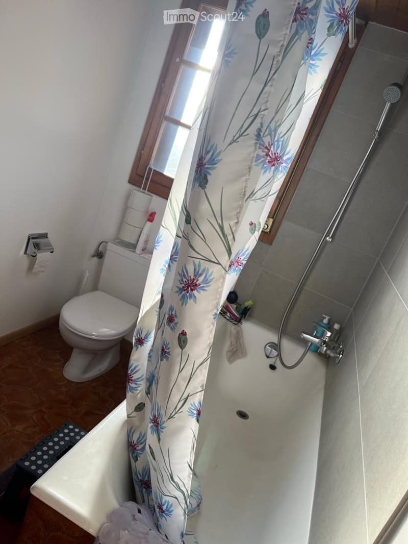 Bathroom with a bathtub, toilet, shower curtain, and a window above the tub. The floor is wooden, and there are towels and toiletries visible.