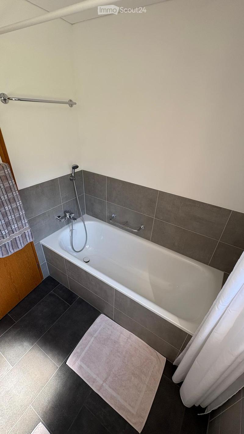 Modern bathroom with a white bathtub, grey tiles, a showerhead, a towel, and a curtain.
