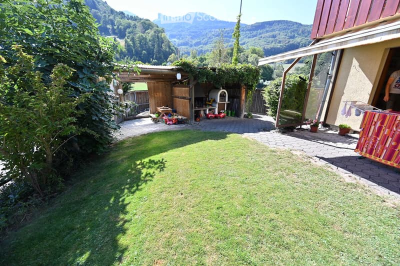 A well-maintained garden with a grassy lawn, a wooden pergola, a pizza oven, and a small wooden shed. The area is surrounded by trees and offers a scenic mountain view in the background.