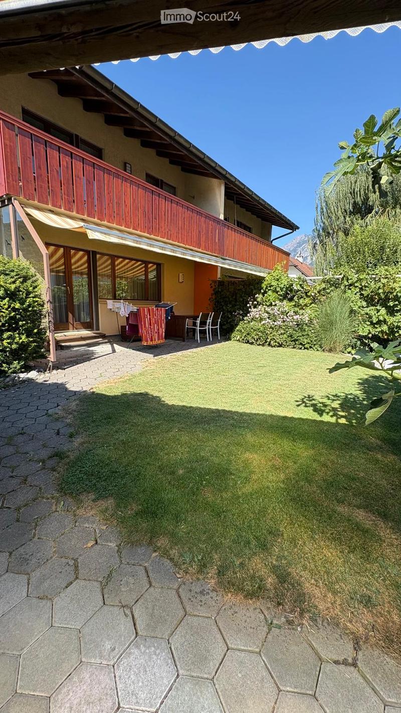 Garden area with a well-maintained lawn, stone patio, outdoor furniture, and a view of a house with a balcony and wooden railing.