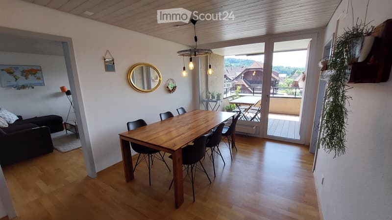 Bright living and dining area with wooden flooring, a large wooden dining table with chairs, a sofa, a hanging pendant light, and a balcony with outdoor furniture visible through large glass doors.