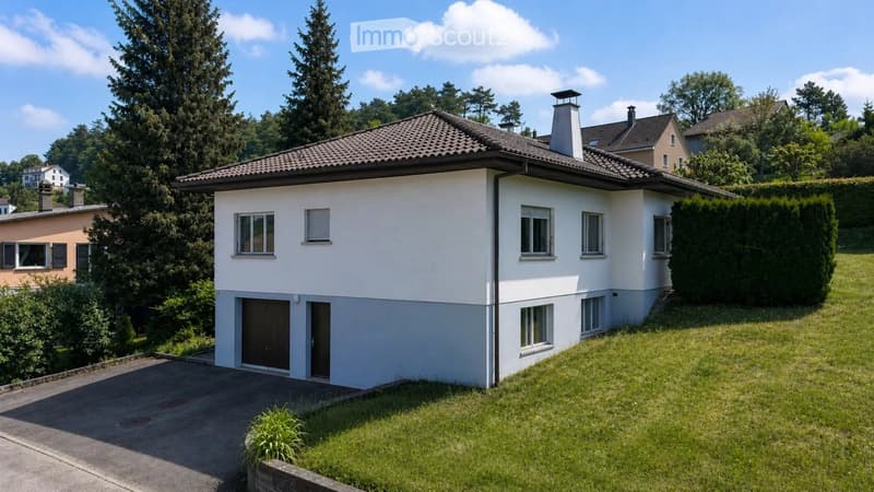 Single-story house with white exterior, tiled roof, garage, and surrounding greenery.