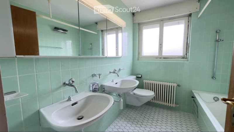 Light green tiled bathroom with two sinks, a toilet, a bathtub, a radiator, and a mirror above the sinks.