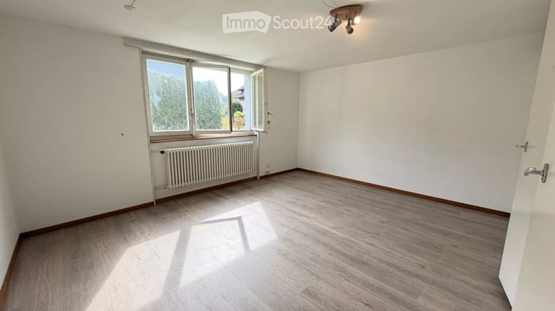 Empty room with wooden flooring, white walls, large window with view, radiator, and ceiling light fixture.