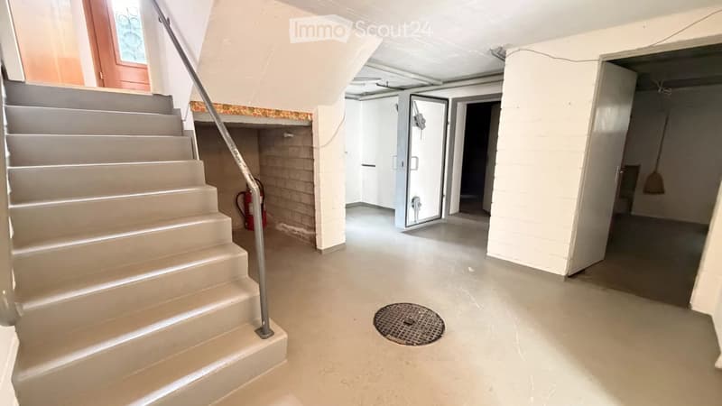 Empty basement hallway with a staircase leading upwards, a fire extinguisher, and a drain cover on the floor.