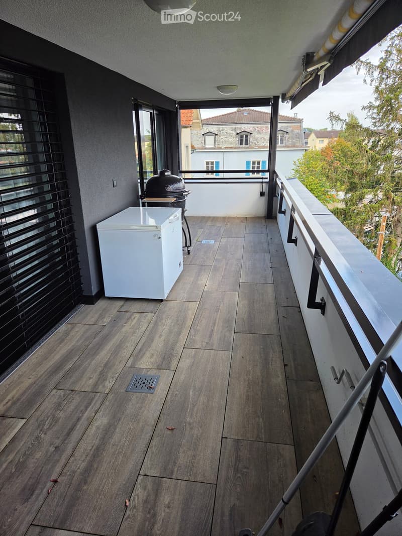 Balcony with wooden flooring, white refrigerator, and grill, overlooking a residential area with trees and houses.