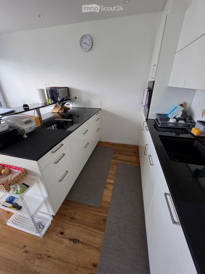 Modern kitchen with white cabinets, black countertops, wooden flooring, and various appliances including a sink, fridge, oven, microwave, toaster, and a clock on the wall.