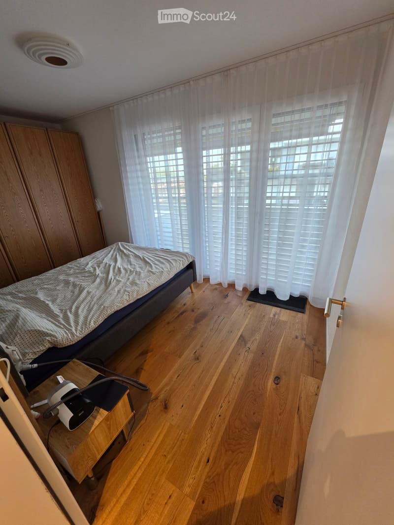 Bedroom with wooden flooring, a bed with a grey duvet, a nightstand with a lamp, and a wardrobe. Large windows with white curtains.