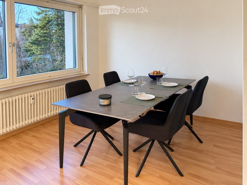 Modern dining room with a sleek black dining table, six black chairs, wine glasses, plates, a bowl, and a candle on the table. Large window with a view of greenery.