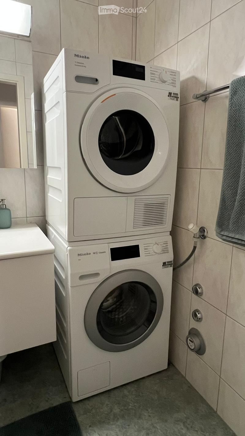 A compact bathroom with a stacked Miele washing machine and dryer unit, a small sink with a soap dispenser, and a towel hanging on the wall.
