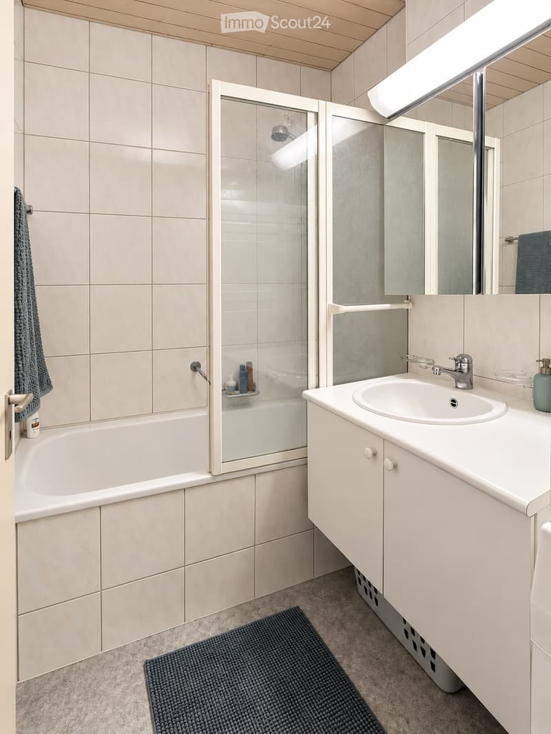Modern bathroom with a bathtub, glass shower stall, sink with cabinet, mirror, and towel. Light-colored tiles and wooden ceiling.
