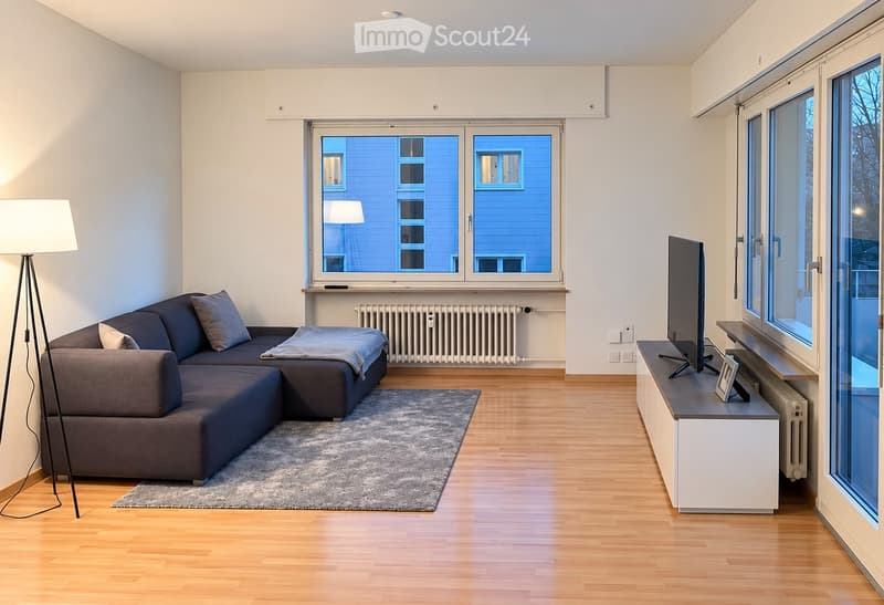 Modern living room with a grey sectional sofa, a coffee table, a floor lamp, a TV stand, and a picture frame on the shelf. The room features large windows with a view of the city and a grey rug on the wooden floor.
