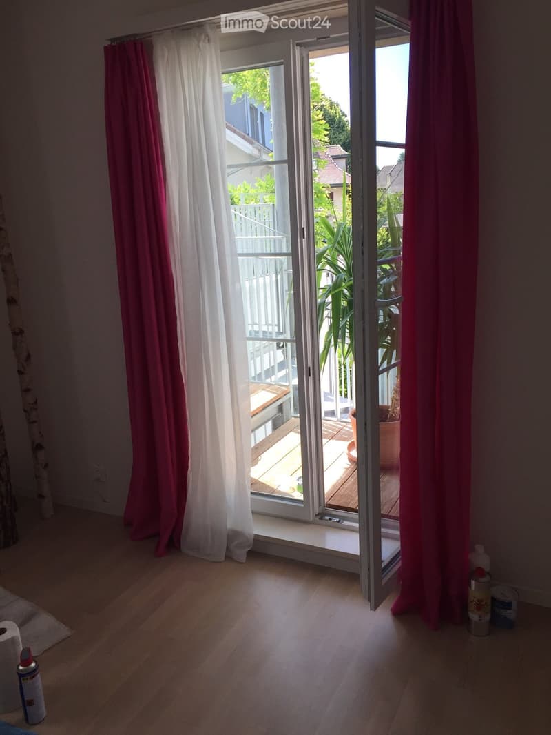 Empty room with wooden flooring, large window with pink and white curtains, and an open sliding door leading to a balcony with wooden decking and potted plants.