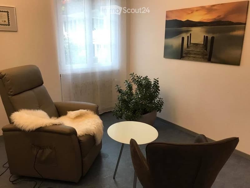 A cozy, well-lit room with a recliner chair, a small round table, a potted plant, and a large painting on the wall.