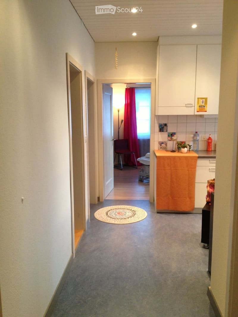 A narrow hallway with carpeted flooring, light-colored walls, and a round decorative rug placed in the center. The hallway leads to an open doorway revealing a room with a red curtain and a chair, while a small kitchen area is visible on the right side wi