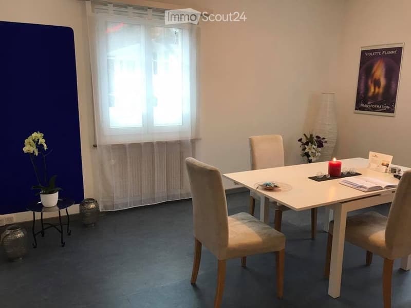 A modern dining area with a white dining table, four beige chairs, a small side table with a flower pot, a decorative vase, and a lit candle on the table. The room features large windows with sheer curtains and a blue accent wall.