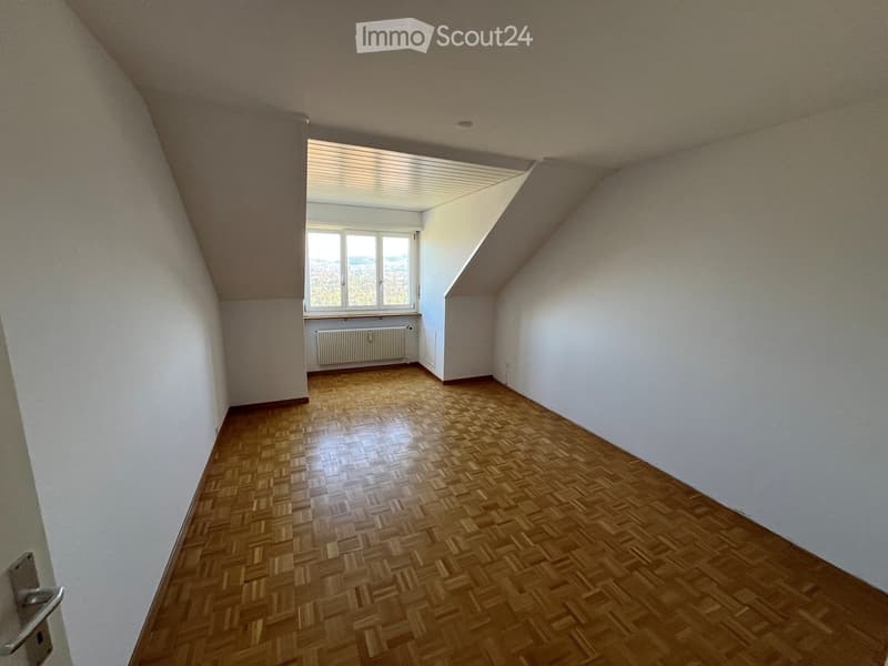 Empty attic room with wooden flooring, white walls, and a large window offering a view. The room features a slanted ceiling and a radiator below the window.