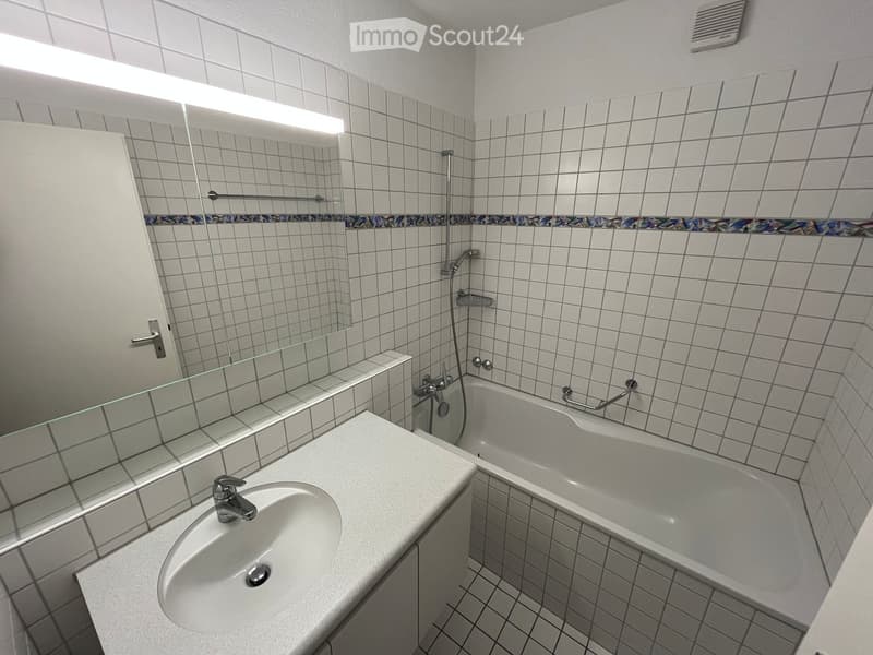 Modern bathroom with white tiled walls and floor, featuring a sink, bathtub, and large mirror with built-in lighting.