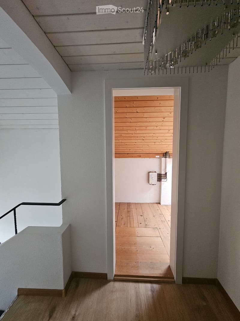 Modern hallway with wooden flooring, white walls, and a staircase leading upwards. A small room with wooden paneling is visible at the end of the hallway.