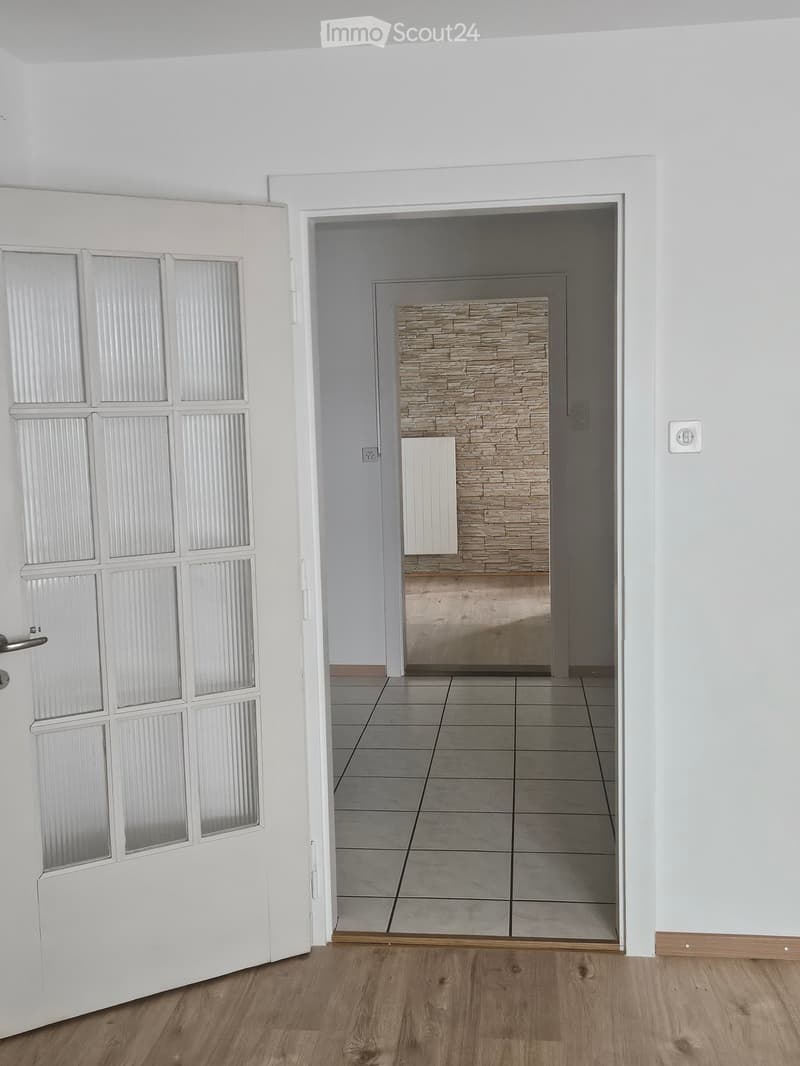Empty hallway with wooden flooring, white walls, and a door leading to another room with a tiled floor and a stone fireplace.