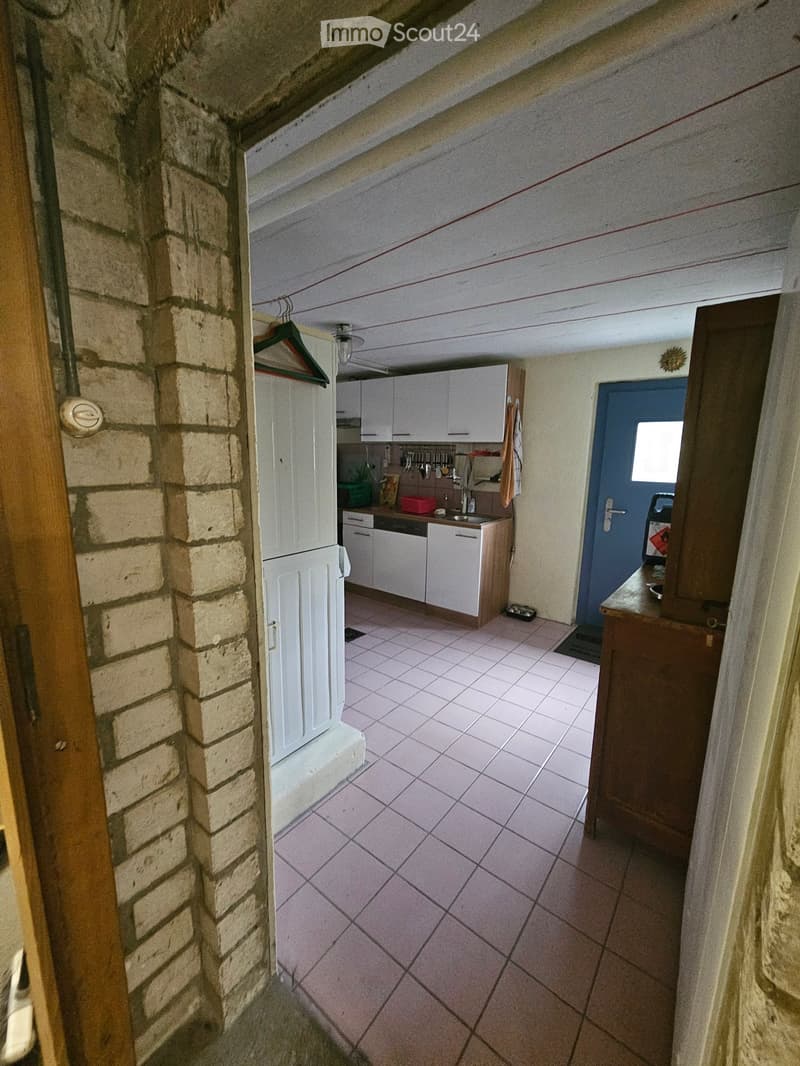 A narrow hallway with a tiled floor, leading to a kitchen area with white cabinets, a sink, and various kitchen utensils. A wooden door is visible on the left, and a blue door is on the right.