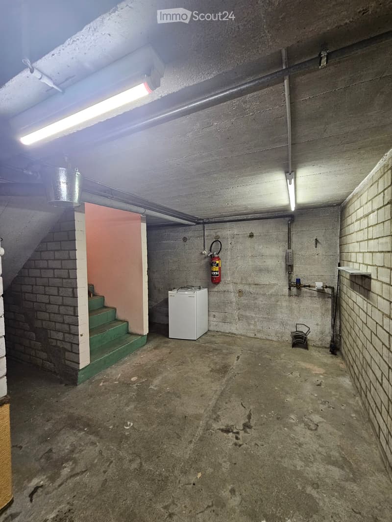 Empty basement with concrete walls and floor, green-painted staircase, white mini-fridge, fire extinguisher, and exposed pipes.