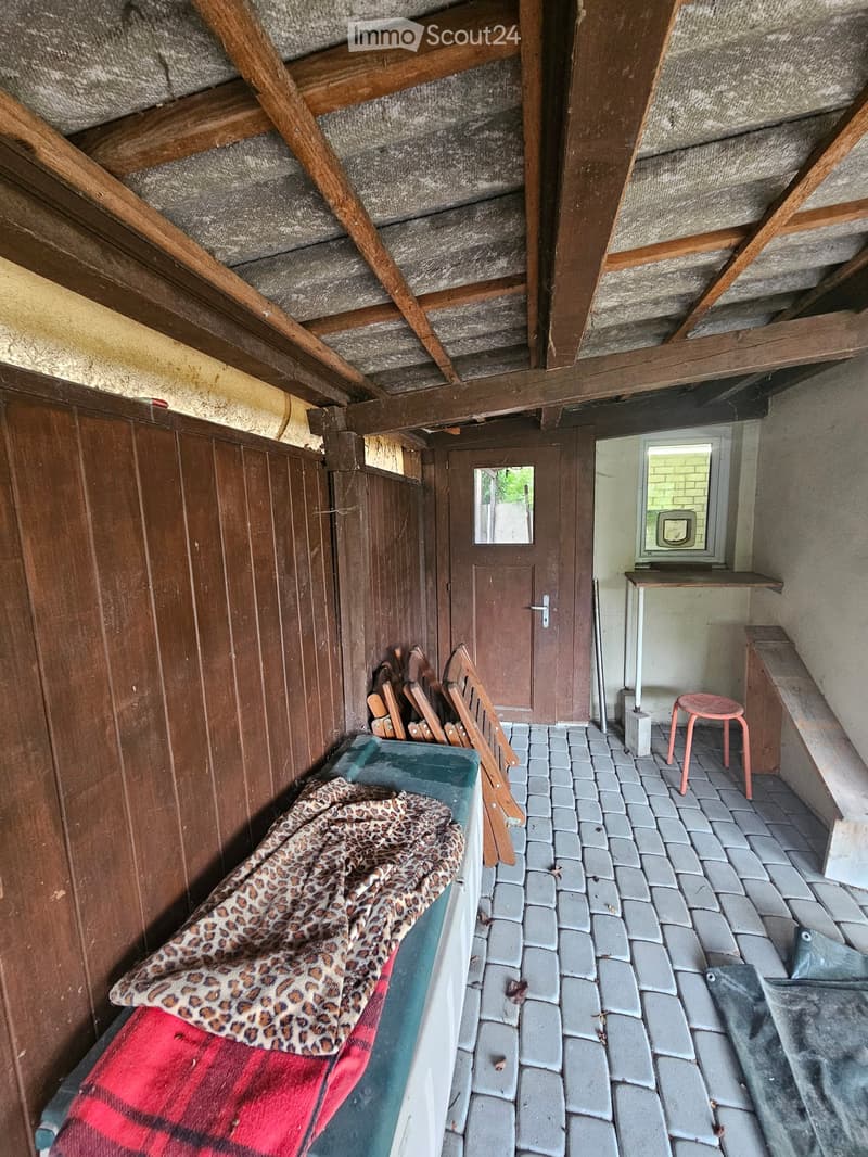A small, wooden-paneled room with a high, slanted wooden ceiling. Features include a small table, a pink stool, a bench, a door with a mirror, and a bed with a leopard-print blanket.