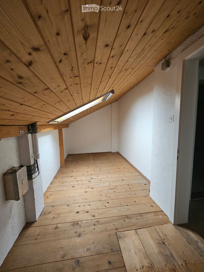 Empty room with wooden floor and slanted wooden ceiling, white walls, and a small light fixture on the ceiling.