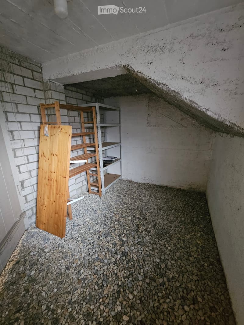 Empty basement space with concrete walls and ceiling, covered in small rocks, with a wooden shelf and a metal shelving unit against the wall.