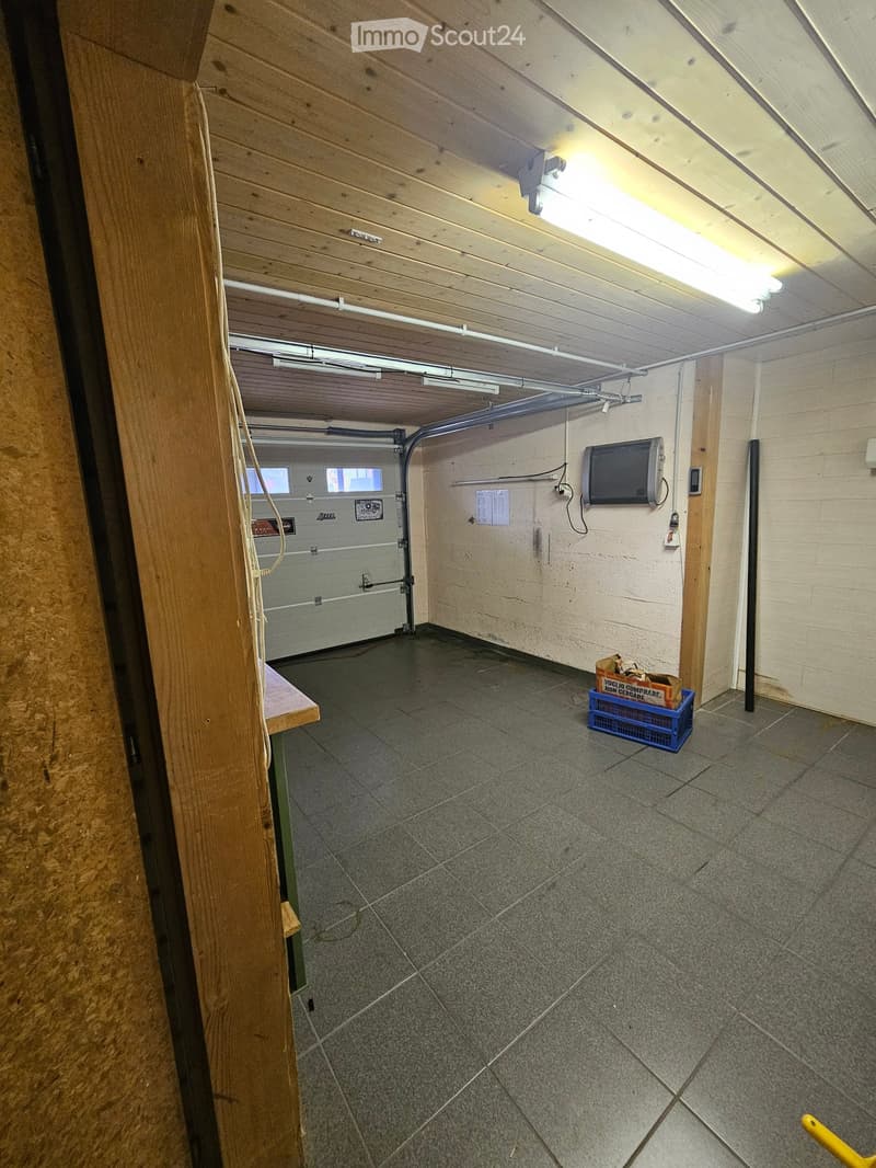 Empty garage with tiled floor, wooden beams, fluorescent lighting, and a partially visible television on the wall.
