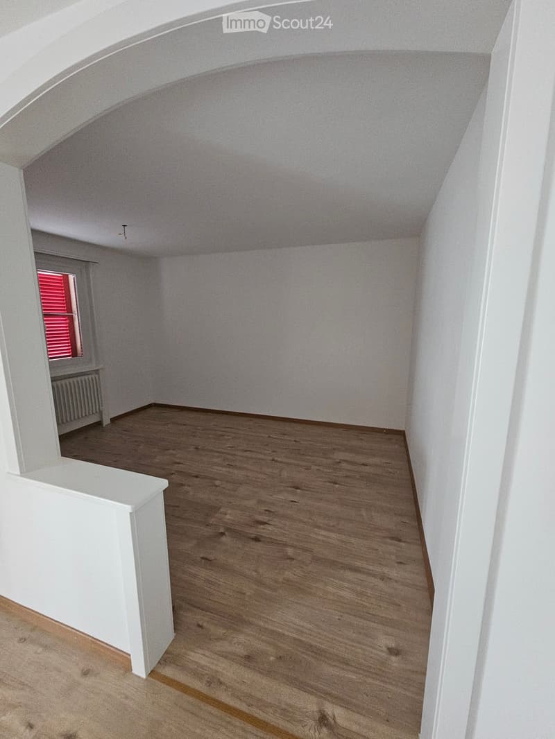 Empty attic room with wooden flooring, white walls, and a slanted ceiling. A window with red shutters is visible in the background.