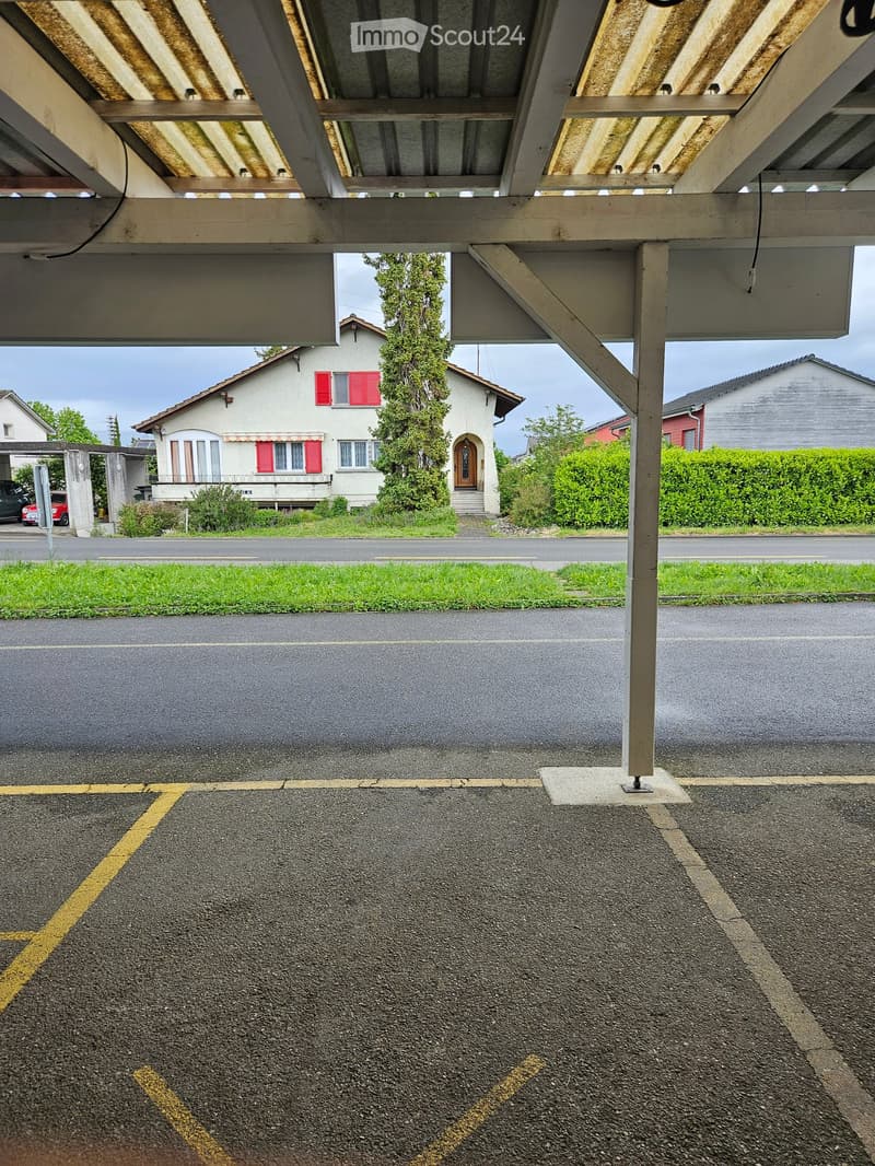 Residential building with a covered parking area, visible parking spaces, and a small garden area.