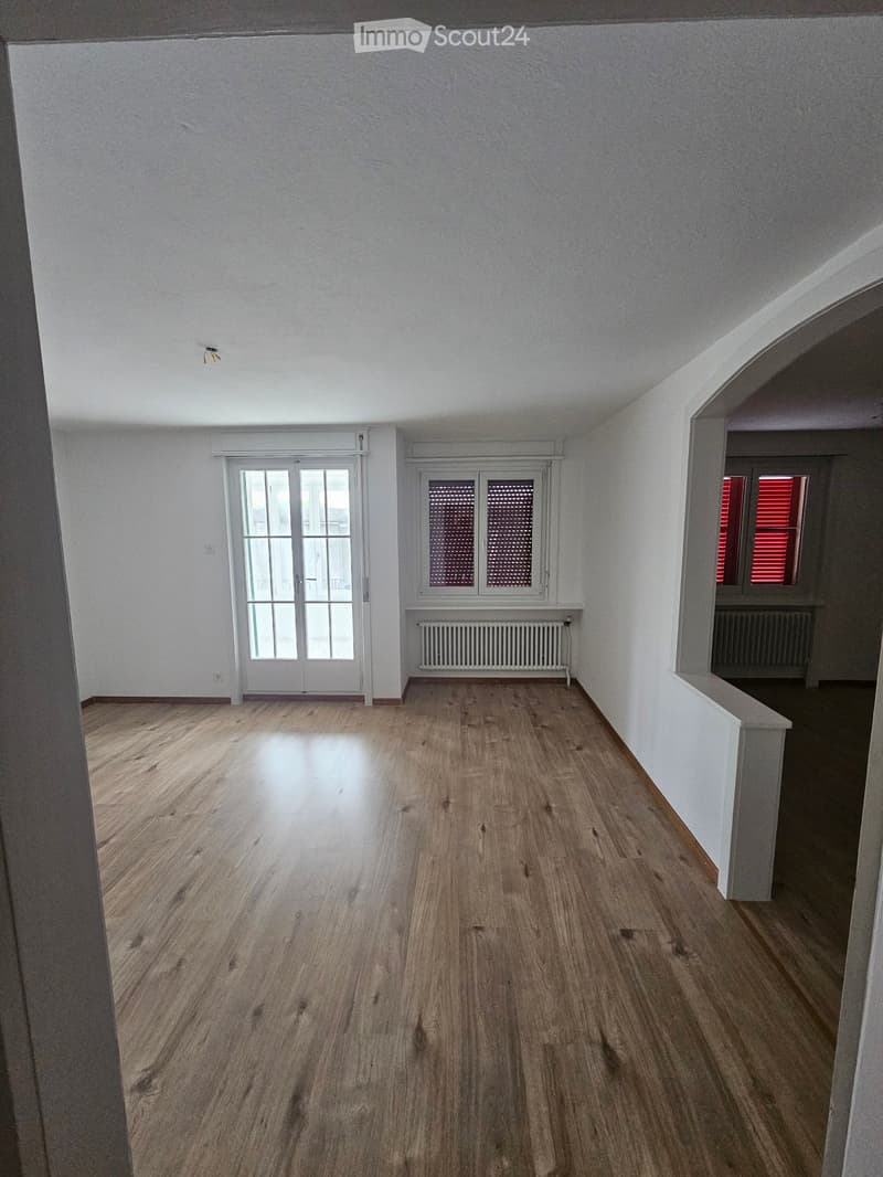 Empty room with wooden flooring, white walls, and large windows with blinds. Radiators are visible under the windows.