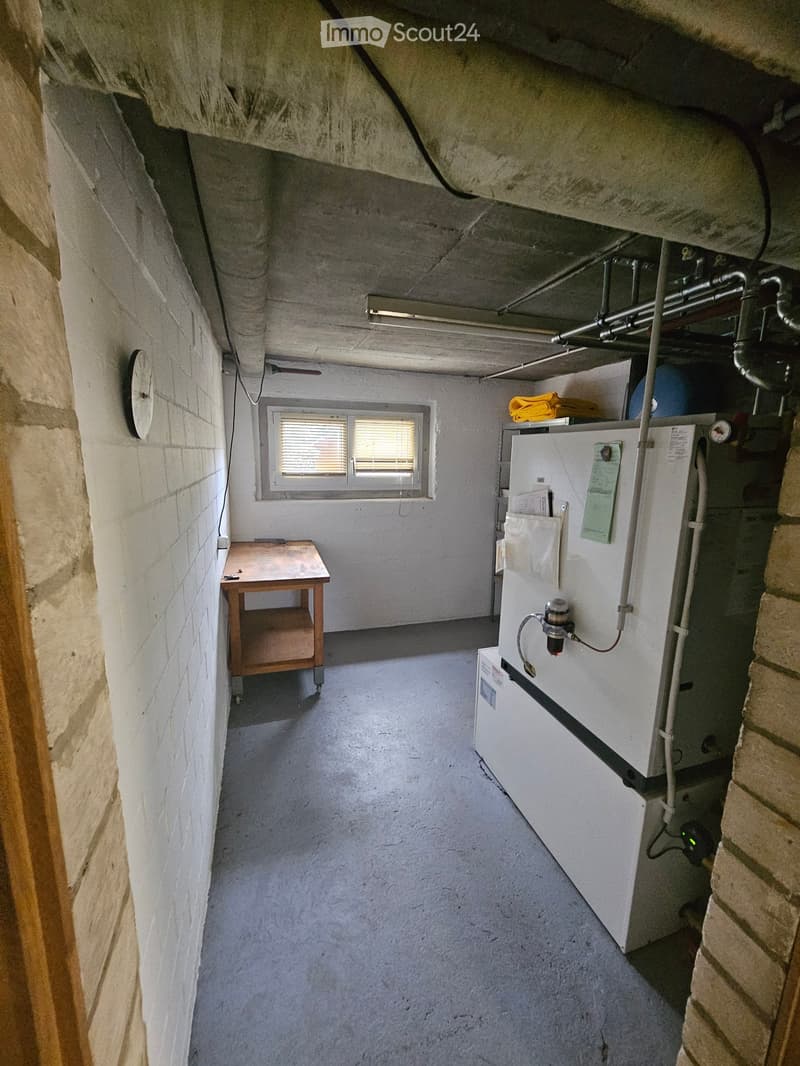 Utility room with a small table, refrigerator, and various pipes and equipment. The room has exposed brick walls and a concrete floor.