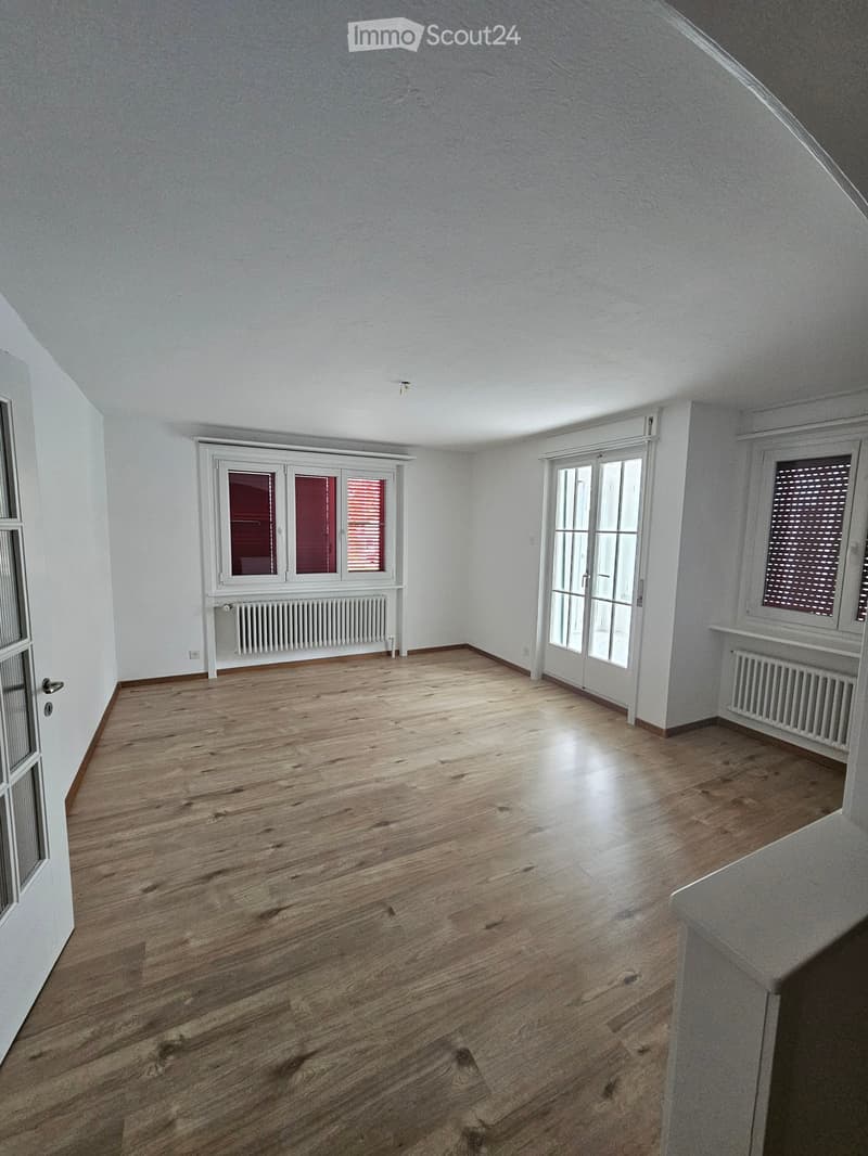 Empty room with wooden flooring, white walls, and large windows with blinds. Radiators are visible beneath the windows.