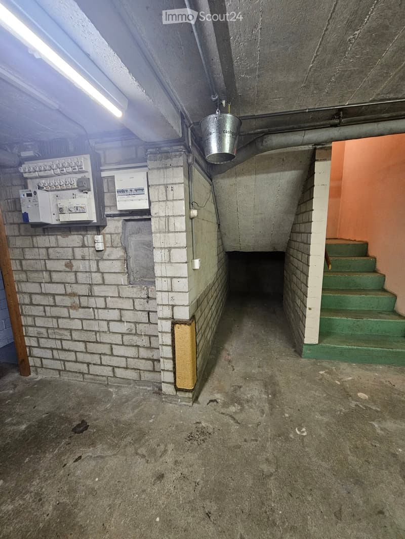 Empty basement corridor with concrete floor, brick walls, electrical panels, green stairs leading up, and a hanging bucket.